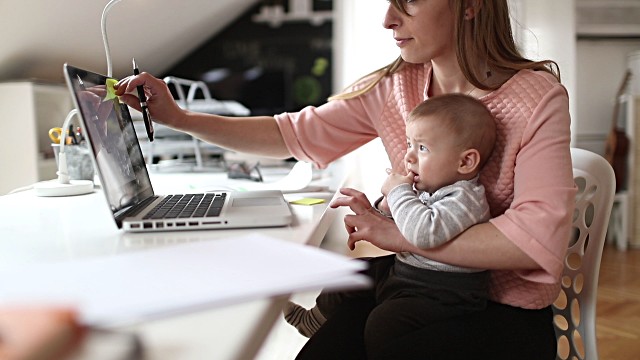 A woman holds a baby in her arms as she works on her laptop at her home