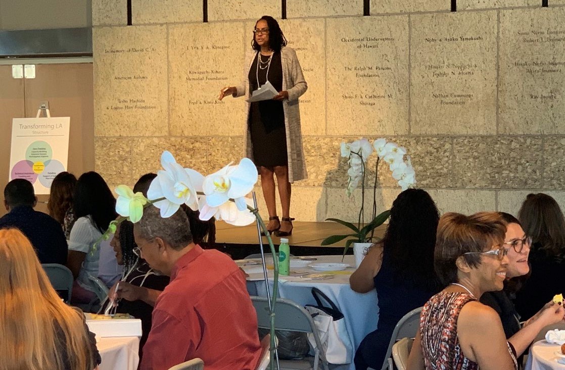 Director of Programs Sheri Dunn Berry stands on stage at the Japanese American National Museum and addresses nonprofit and LA County folks seated at tables with orchid center pieces. 